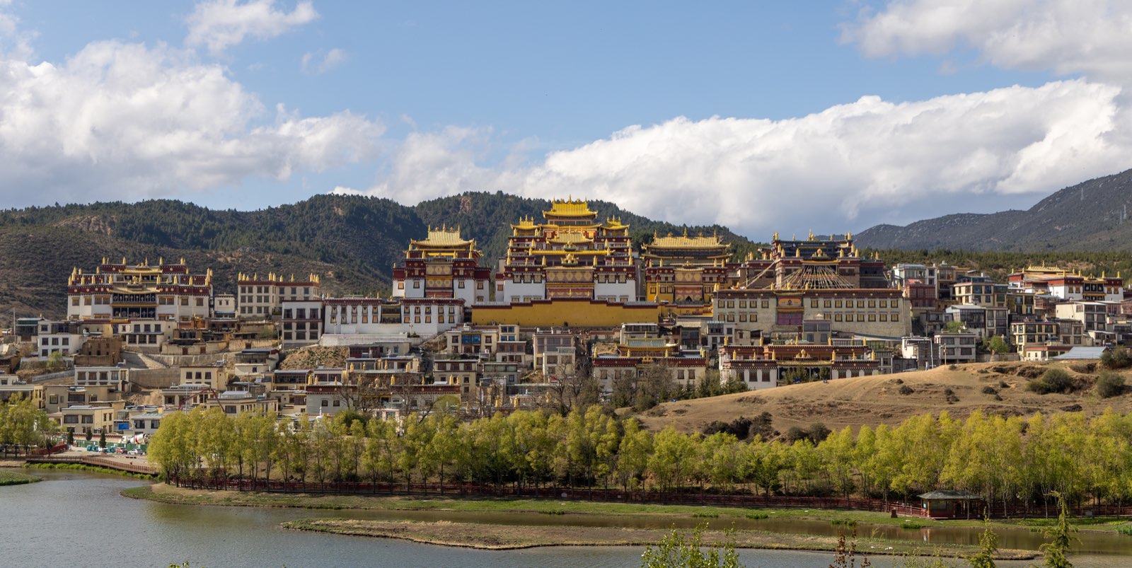 松赞林寺全景：金顶红墙的寺院建筑群沿山坡错落展开，前方一汪湖水倒映蓝天白云，背后是连绵青山